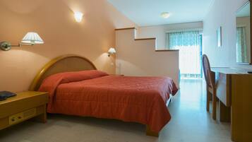 A bedroom with a bed, bedside table, lamp, desk, chair, and a view of a balcony.