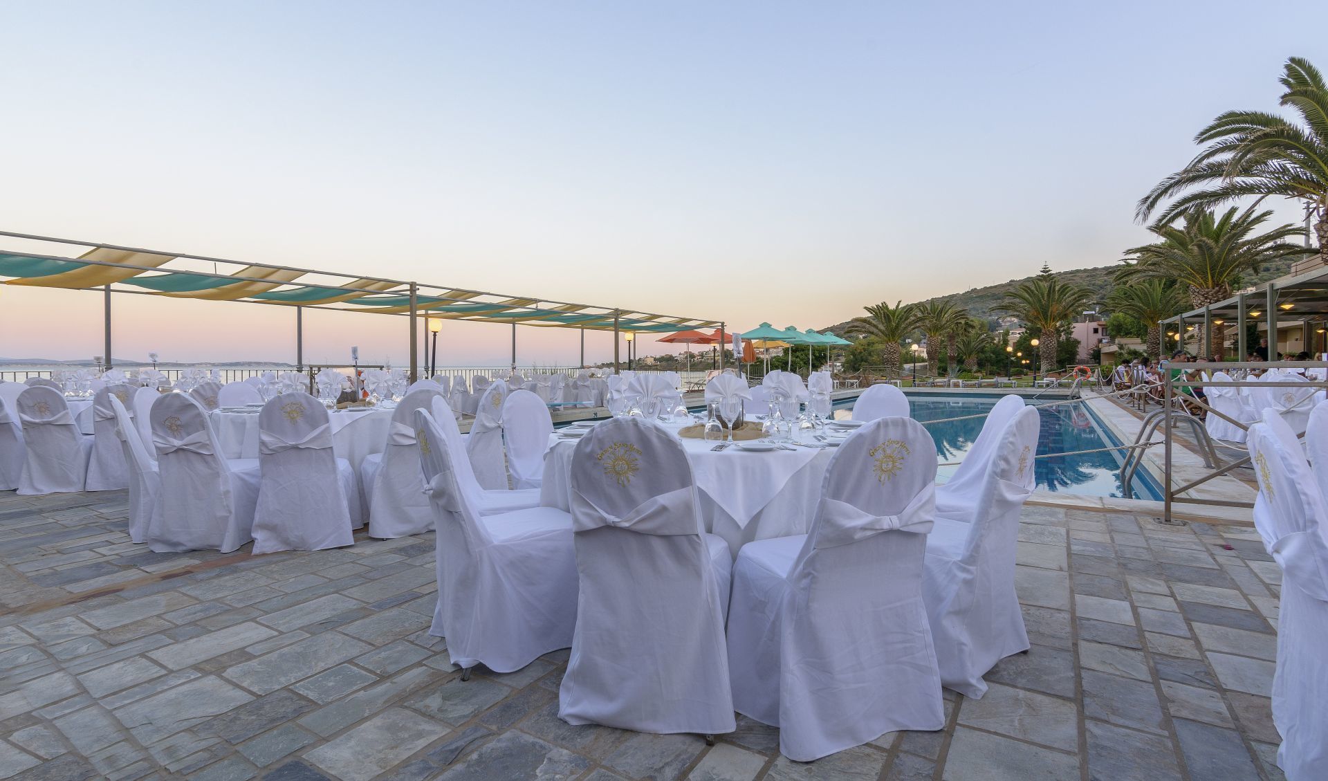 An outdoor dining area with white tablecloths and chairs, a pool, and a view of the sea.