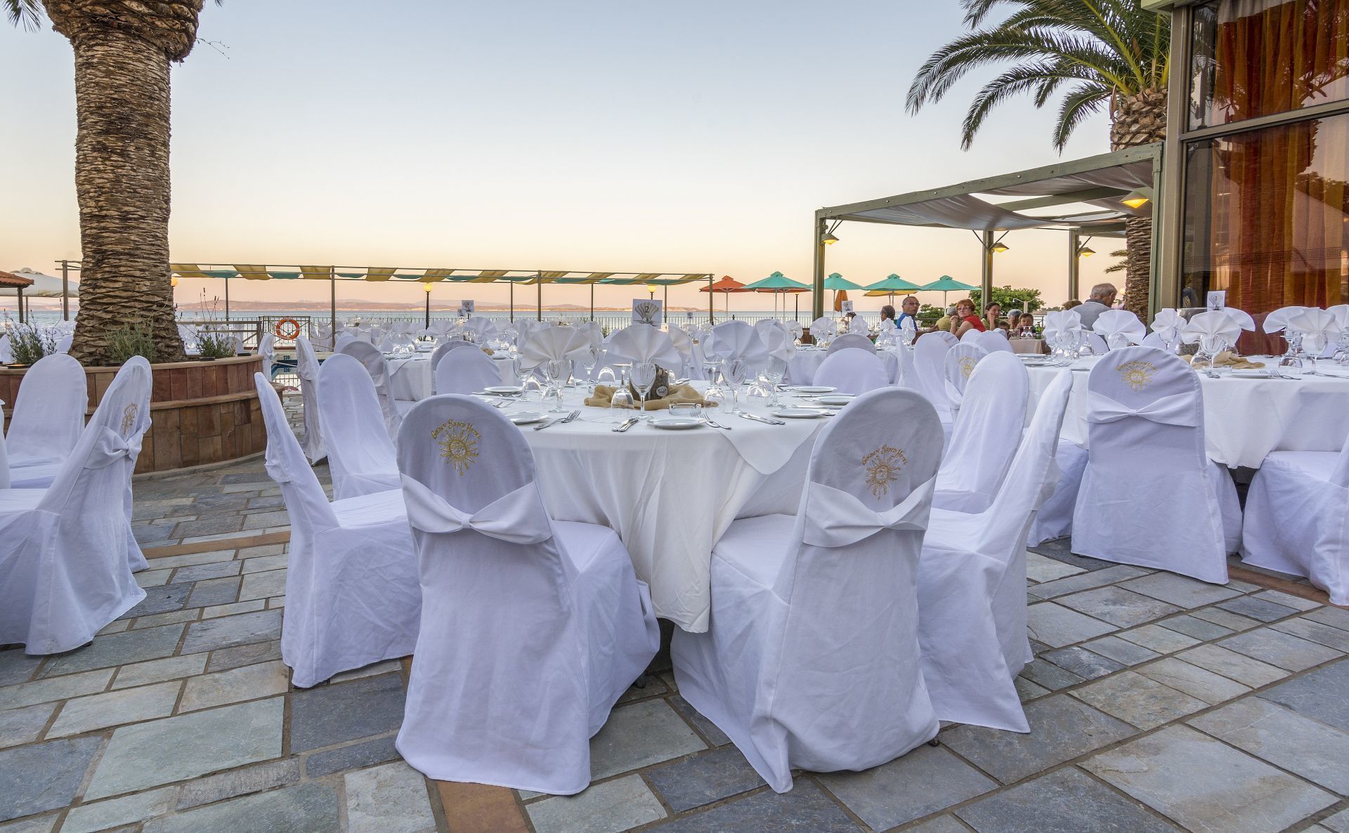An outdoor dining area with round tables set for a formal event, covered with white tablecloths and chairs with white covers.
