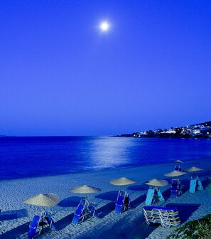 Una playa con filas de sillas azules y sombrillas de paja bajo un cielo nocturno con luna visible.