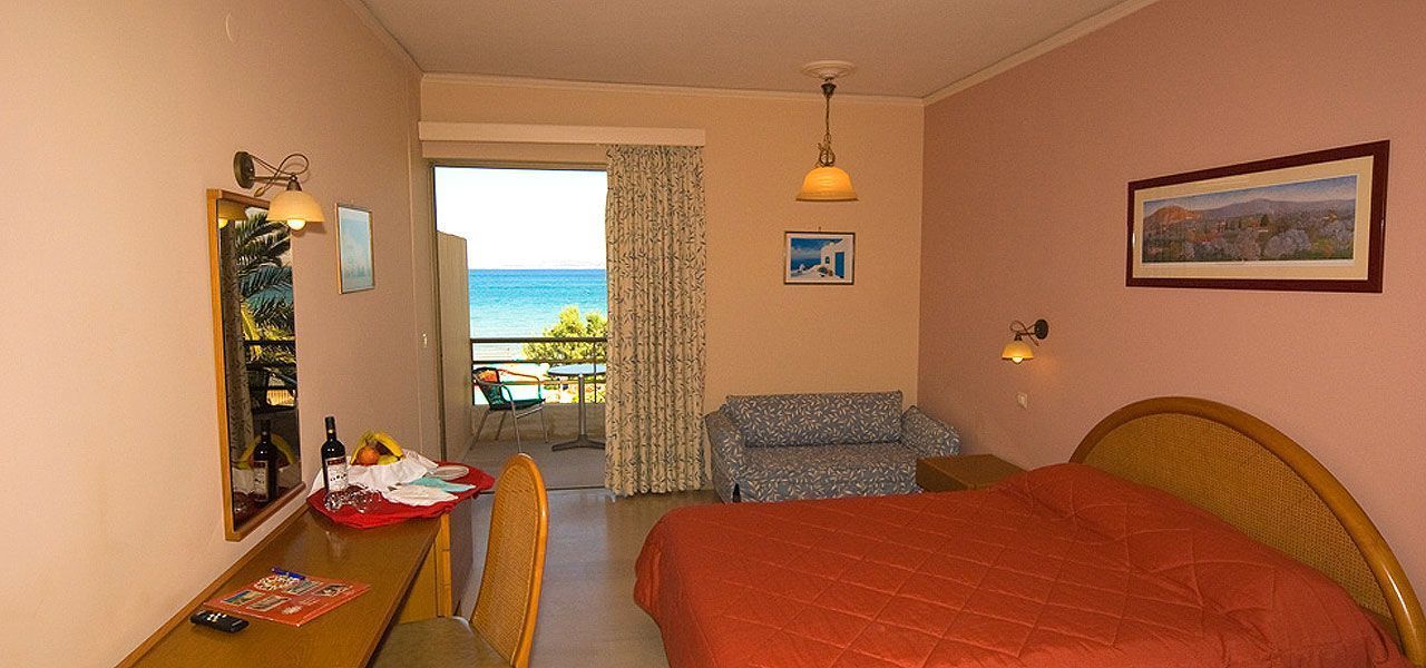 Une chambre d’hôtel avec un lit, un bureau et une chaise, une petite table recouverte d’une nappe rouge, une chaise, un canapé, une fenêtre donnant sur la mer et un tableau accroché au mur.