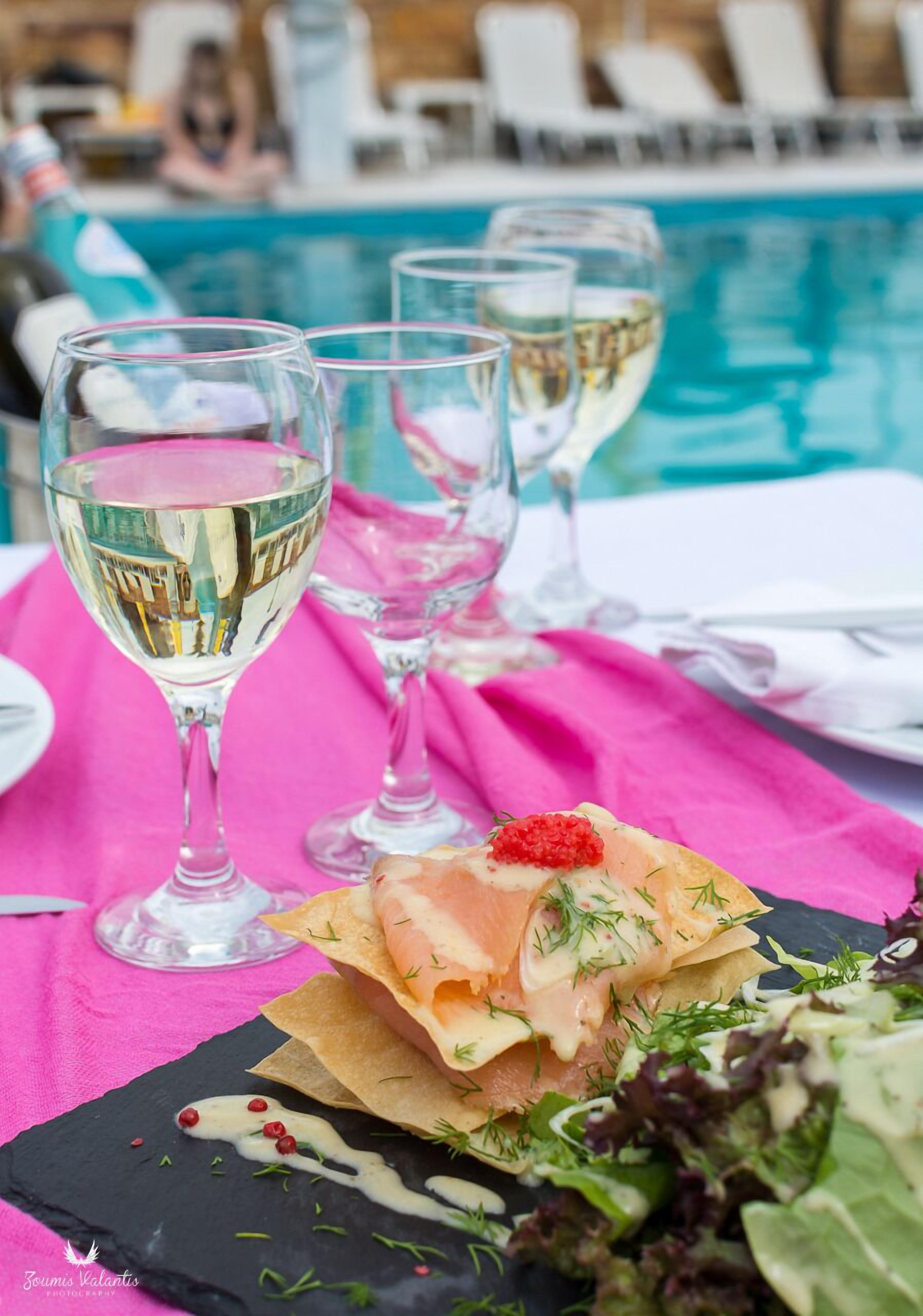 A dining table with a pink tablecloth, three wine glasses, a dish with salmon and greens, and a pool in the background.