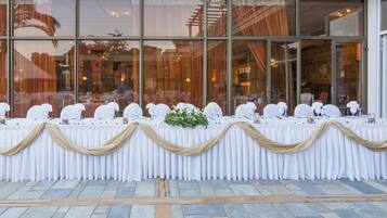 Una mesa de banquete dispuesta con manteles blancos y cortinas doradas, ubicada frente a un edificio con grandes ventanales.