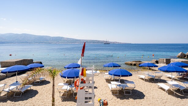 A beach with blue umbrellas, white lounge chairs, and a lifebuoy. - Capo Peloro Resort - Adults Only (Messina)