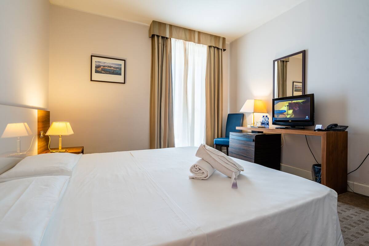 A hotel room with a neatly made bed, a desk with a computer, a chair, and a television.