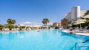 A large outdoor swimming pool with lounge chairs and umbrellas, surrounded by palm trees and buildings. - Capo Peloro Resort - Adults Only (Messina)