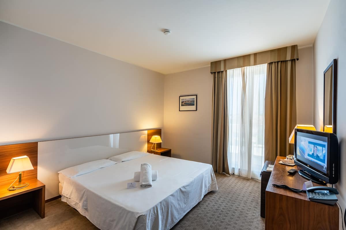 A hotel room with a large bed, two bedside tables with lamps, a TV, and a window with curtains.