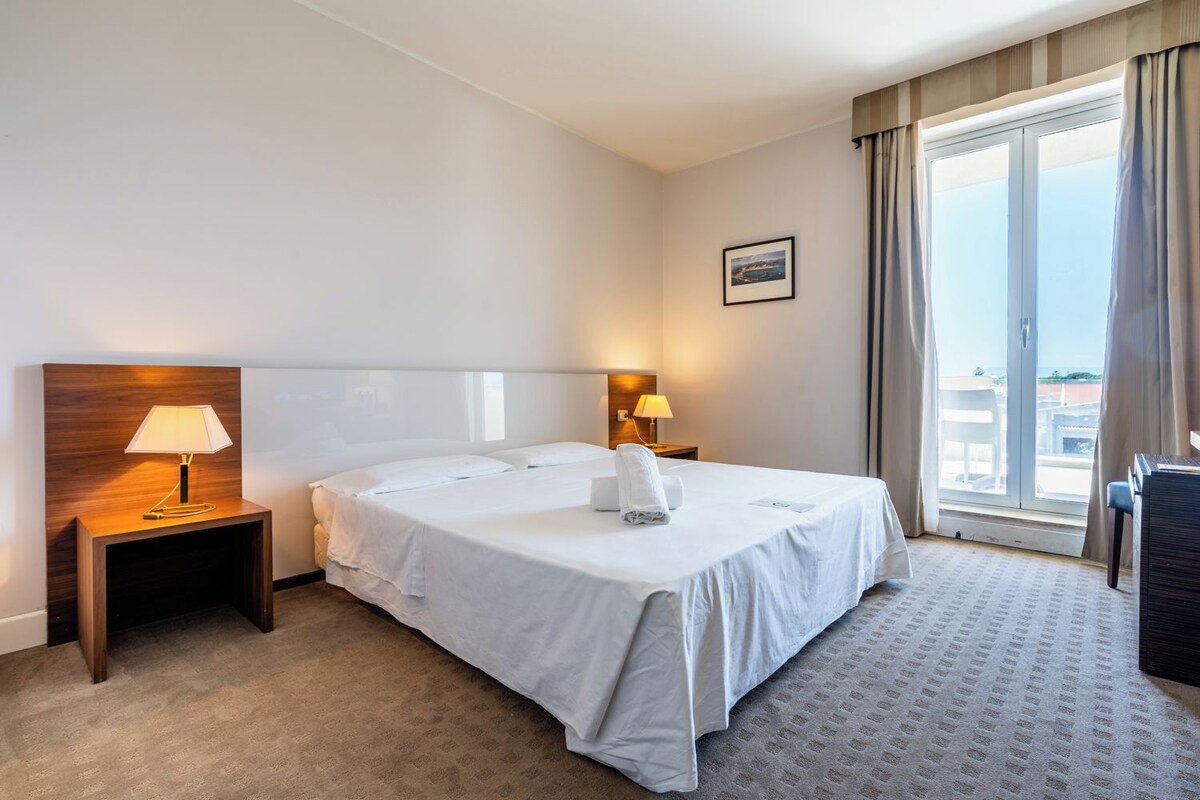 A hotel room with a large bed, two bedside lamps, a headboard, a window with curtains, and a small picture on the wall.