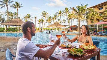 A couple dining by a pool with a tropical setting, including palm trees and a thatched-roof structure in the background.