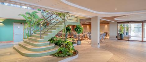 A spacious indoor area with a curved staircase surrounded by greenery, a dining area with tables and chairs, and large windows allowing natural light.