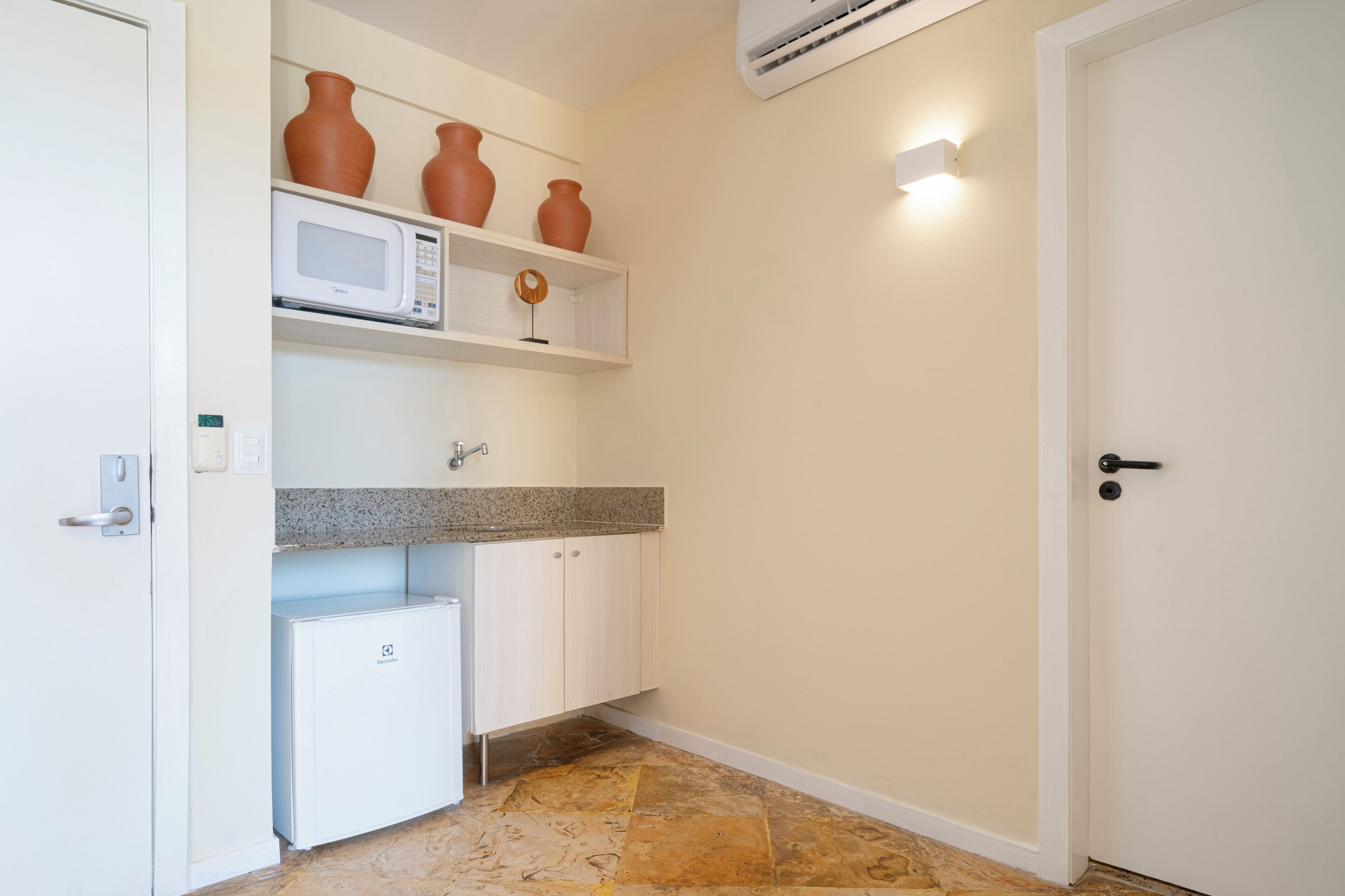 Cocina compacta con armarios blancos, encimera de granito, microondas y refrigerador pequeño. Hay tres macetas de terracota en un estante sobre el microondas.