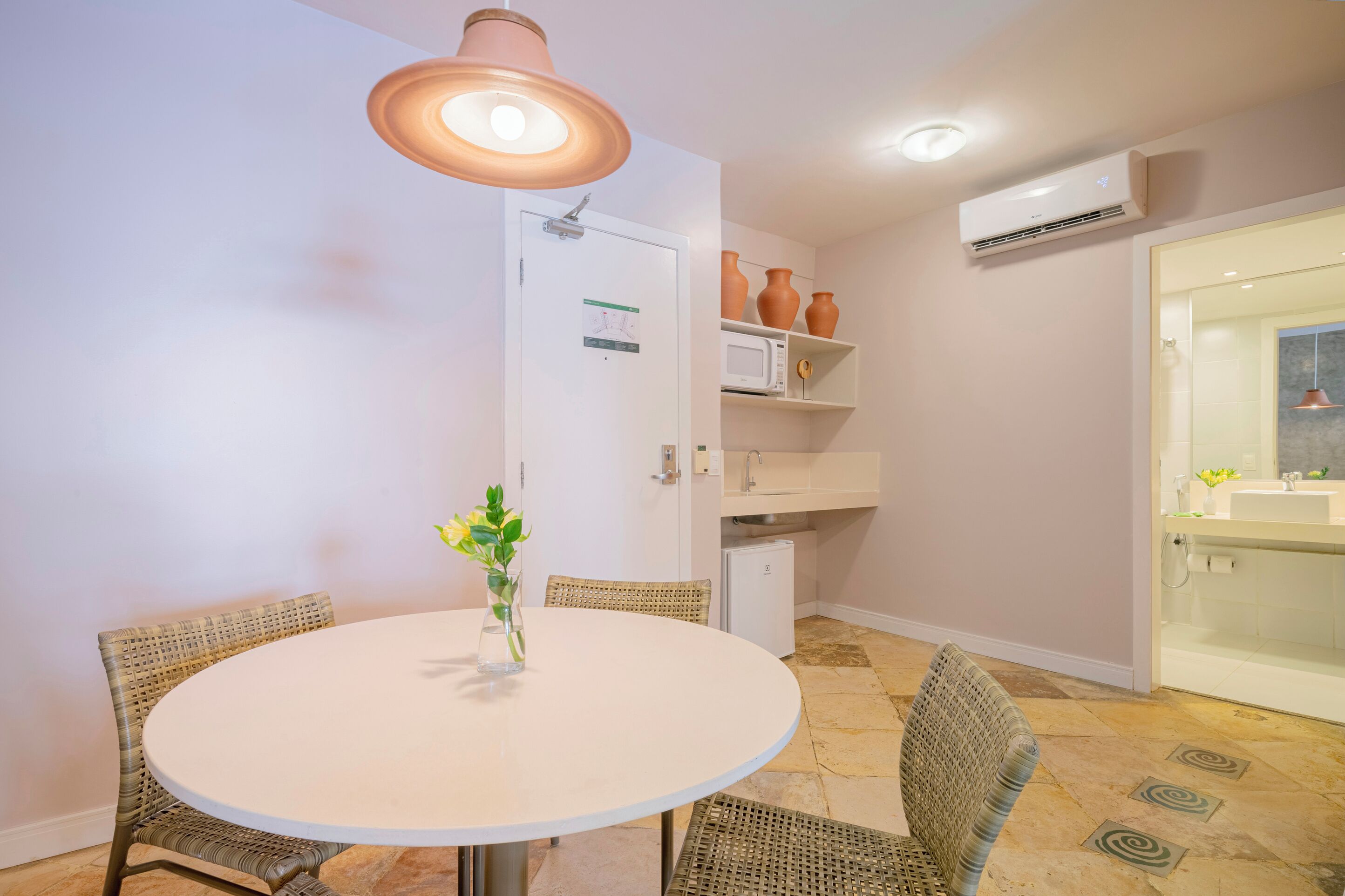 Un comedor con una mesa redonda, sillas de mimbre, una lámpara colgante, una puerta blanca con un calendario, un microondas y un refrigerador pequeño.
