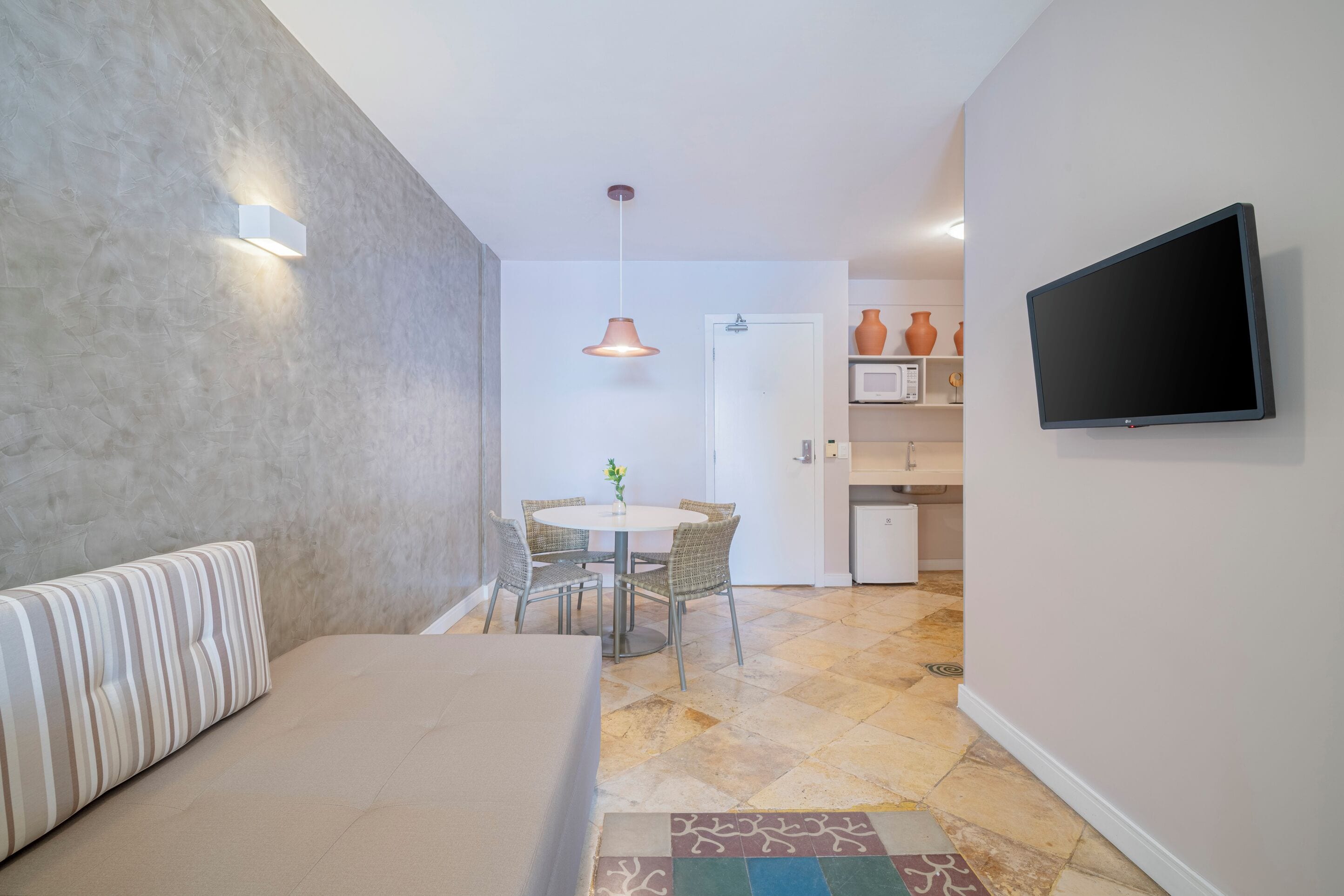 A modern hotel room with a bed, a dining area with a table and chairs, a flat-screen TV mounted on the wall, and a kitchen area with a microwave and cabinets.