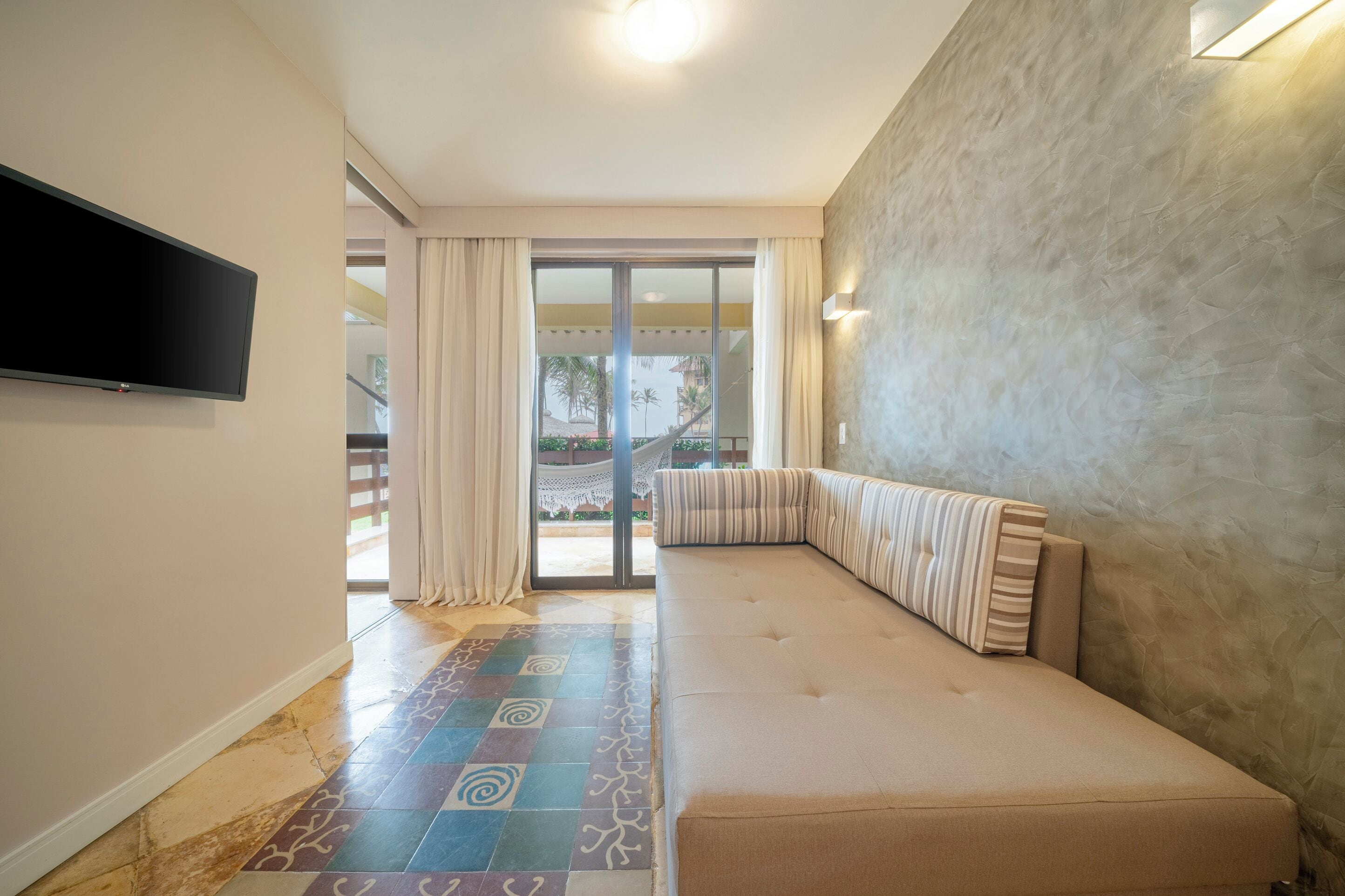 A modern living room with a large sofa, a flat-screen TV mounted on the wall, and a sliding glass door leading to a balcony with a view of palm trees.