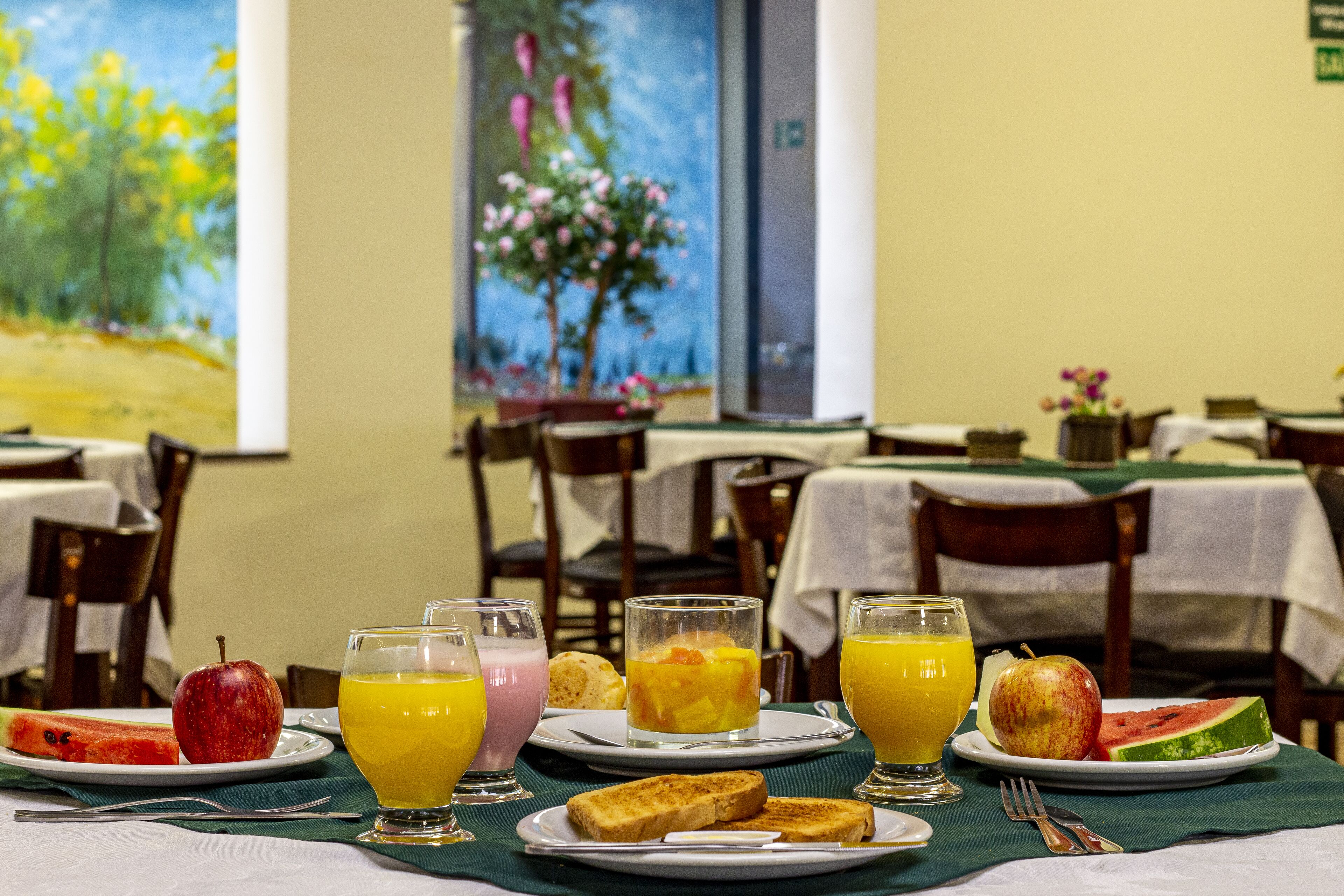 A breakfast setting with plates of fruit, juice, and toast, and a painting on the wall.