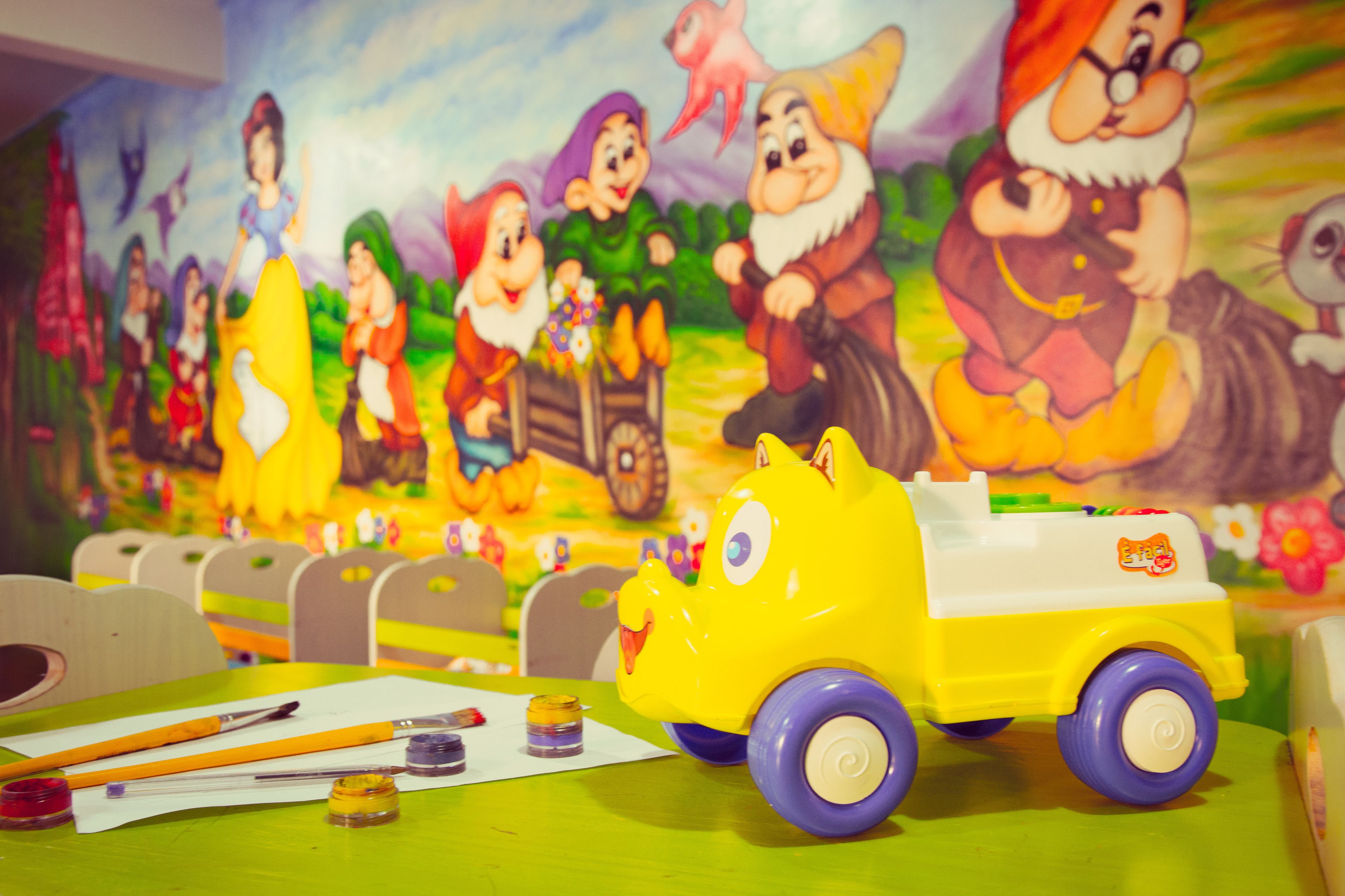 Ein Tisch mit einem Spielzeugauto und Bastelmaterial, eine Wandmalerei mit Zeichentrickfiguren und eine grüne Tischdecke.