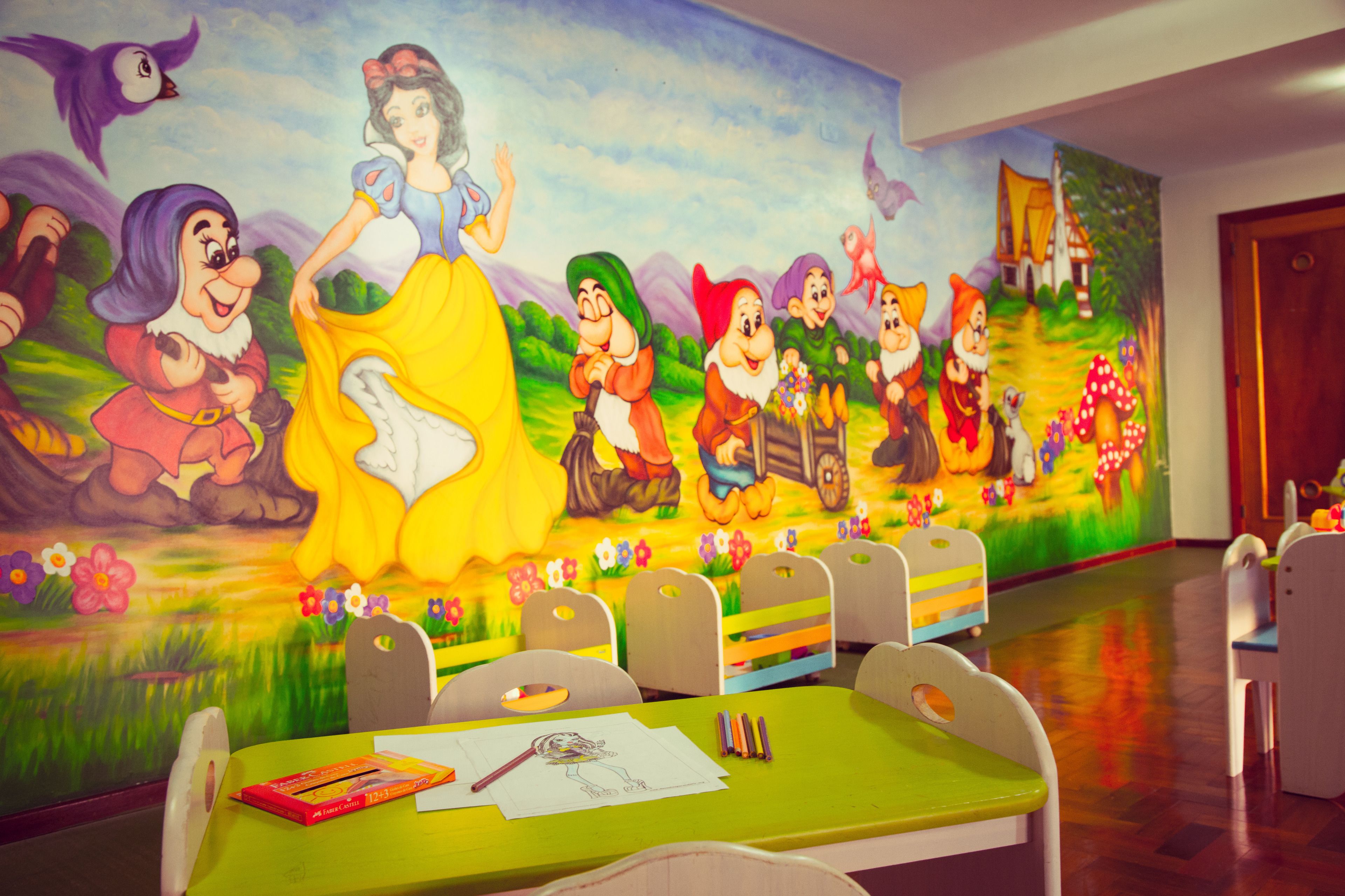 Une chambre d’enfant ornée d’une fresque représentant des personnages de Disney, une table verte avec du matériel de dessin et de peinture, et un plancher en bois.