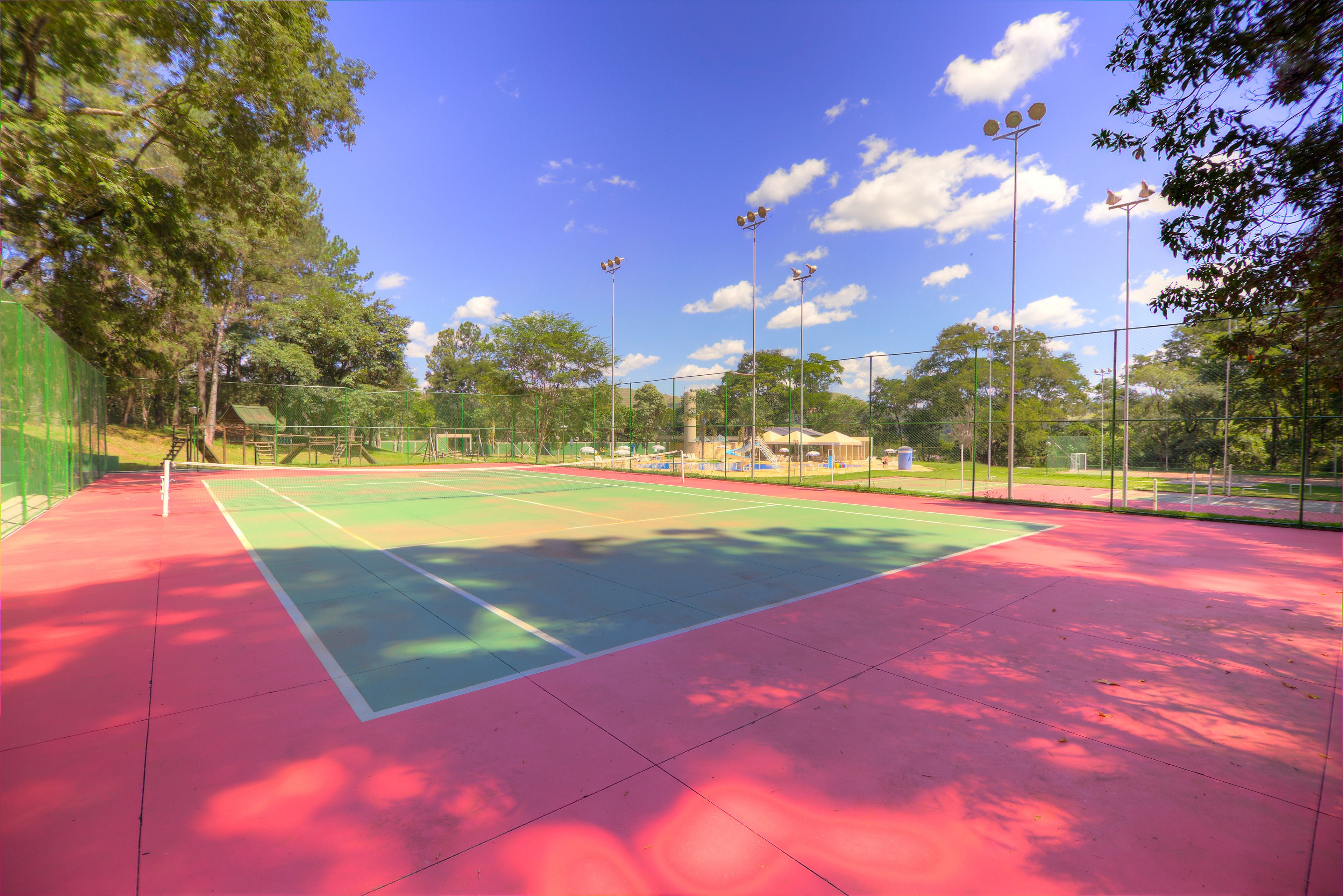 An outdoor tennis court with a green and red surface, surrounded by trees and a fence.