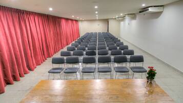Uma sala de conferência com fileiras de cadeiras, uma mesa de madeira e uma cortina vermelha.