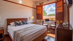 Habitación de hotel con cama, escritorio, silla, televisor y ventana con contraventanas abiertas.