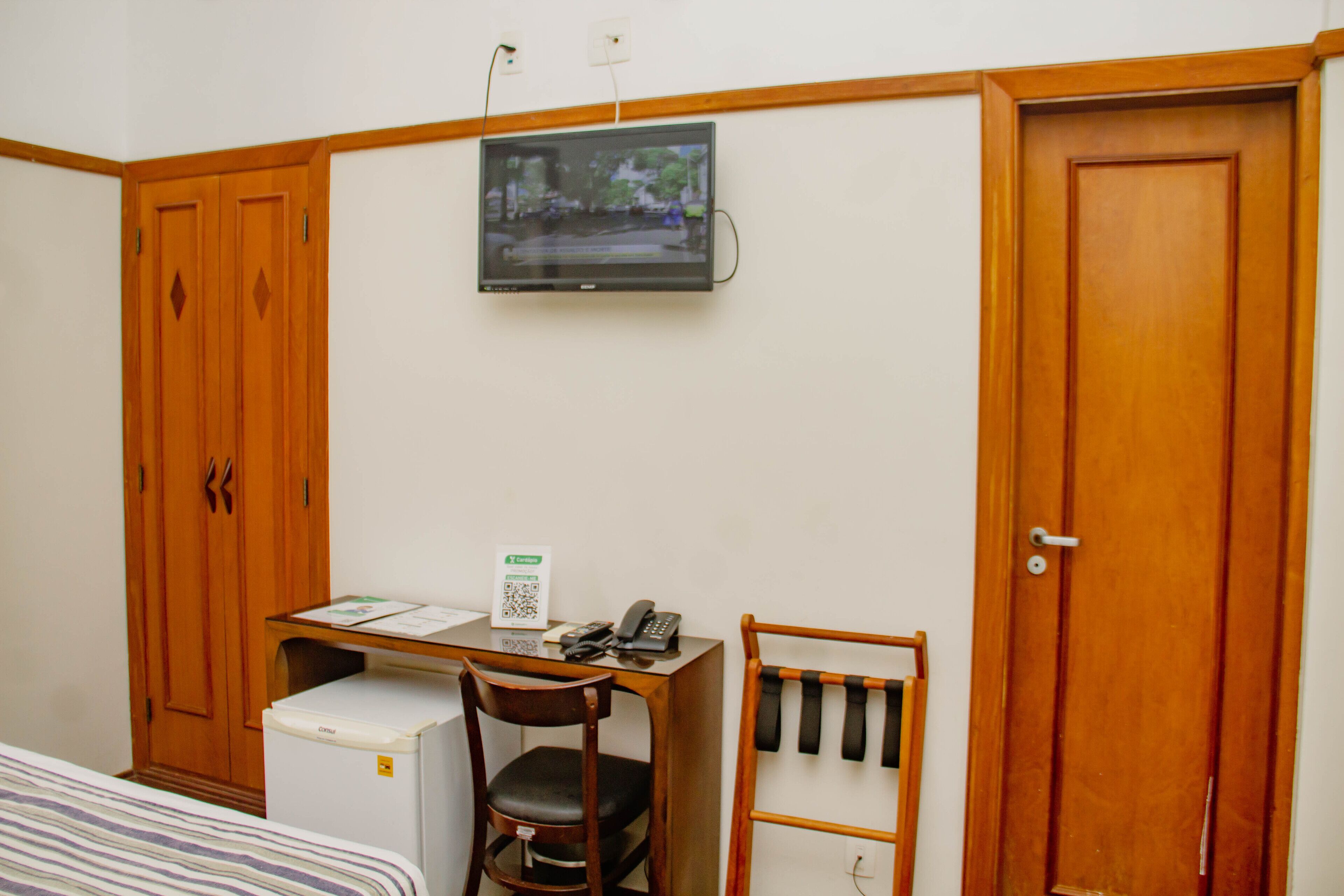 A bedroom with a bed, a desk with a computer, a chair, a small refrigerator, a TV on the wall, and wooden wardrobes.