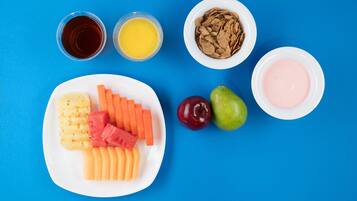Un plato con frutas y verduras cortadas, dos vasos de jugo, un tazón de cereal y un tazón de yogur sobre un fondo azul.