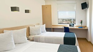 Habitació d'hotel amb dos llits, una taula de treball, una cadira, un televisor i una finestra amb vista a la ciutat.