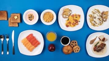 Una mesa azul con una variedad de alimentos para el desayuno, incluyendo tostadas, yogur, cereales, huevos, papas fritas, fruta, jugo, café, croissants y bollería.