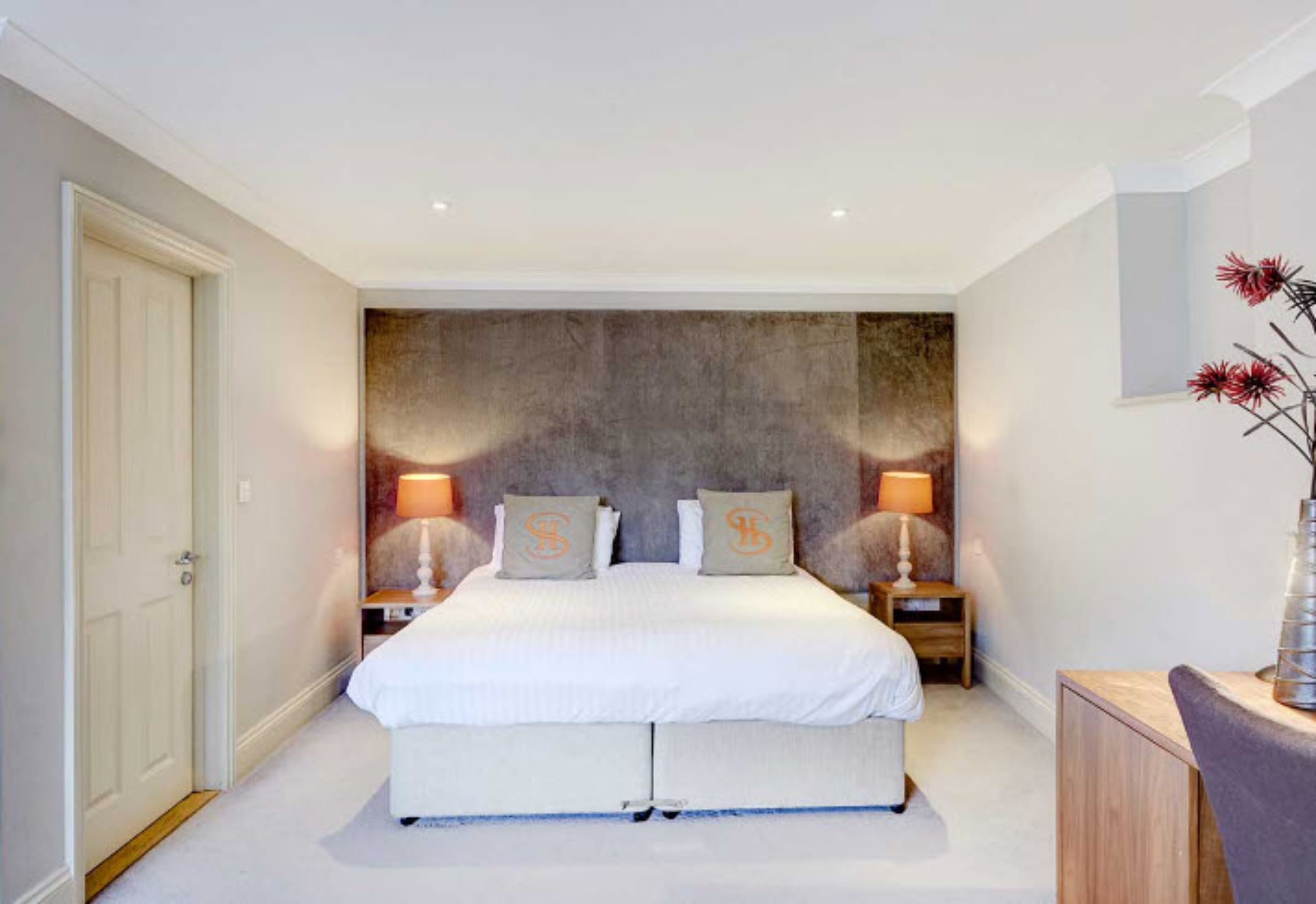 Een slaapkamer met een groot bed, twee nachtkastjes met lampen, een houten dressoir en een deur.