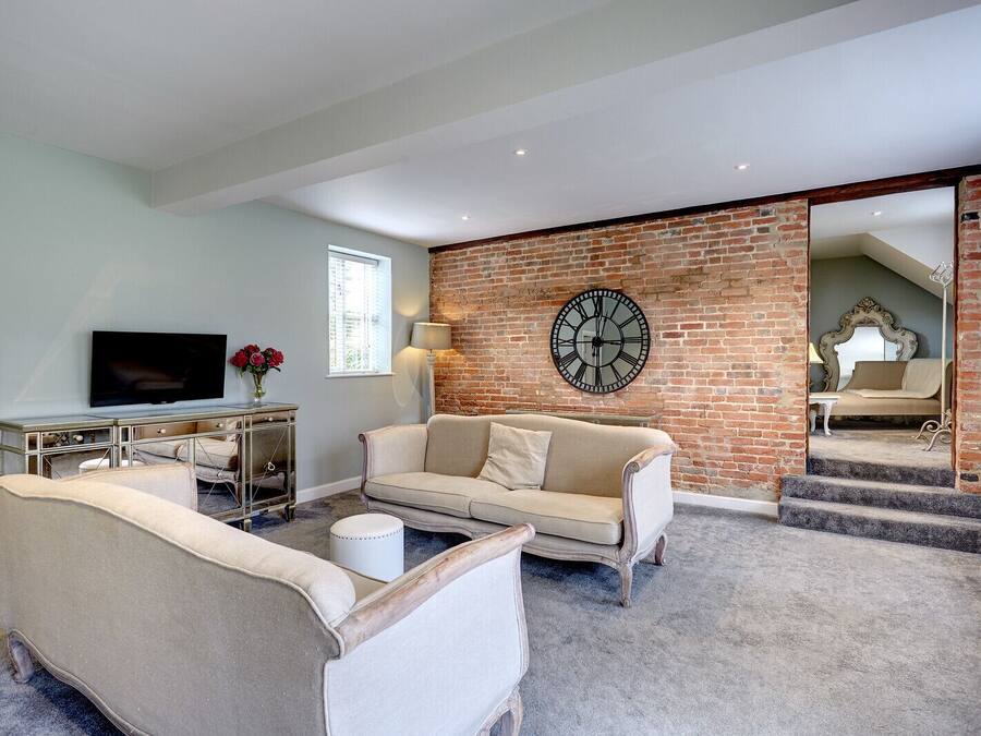 Una sala de estar con pared de ladrillo, un reloj grande, un sofá, un televisor y una escalera que conduce a otra habitación.