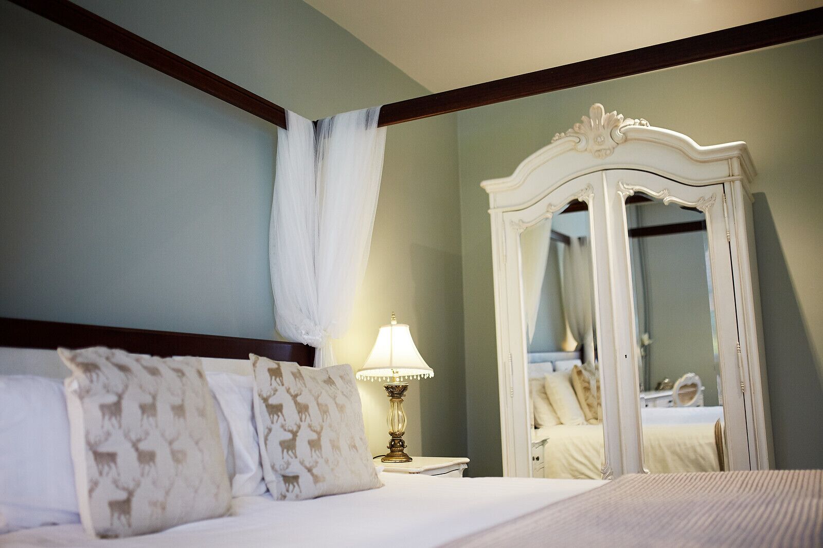 A bedroom with a bed, a nightstand, a lamp, a mirror, and a window with a curtain.
