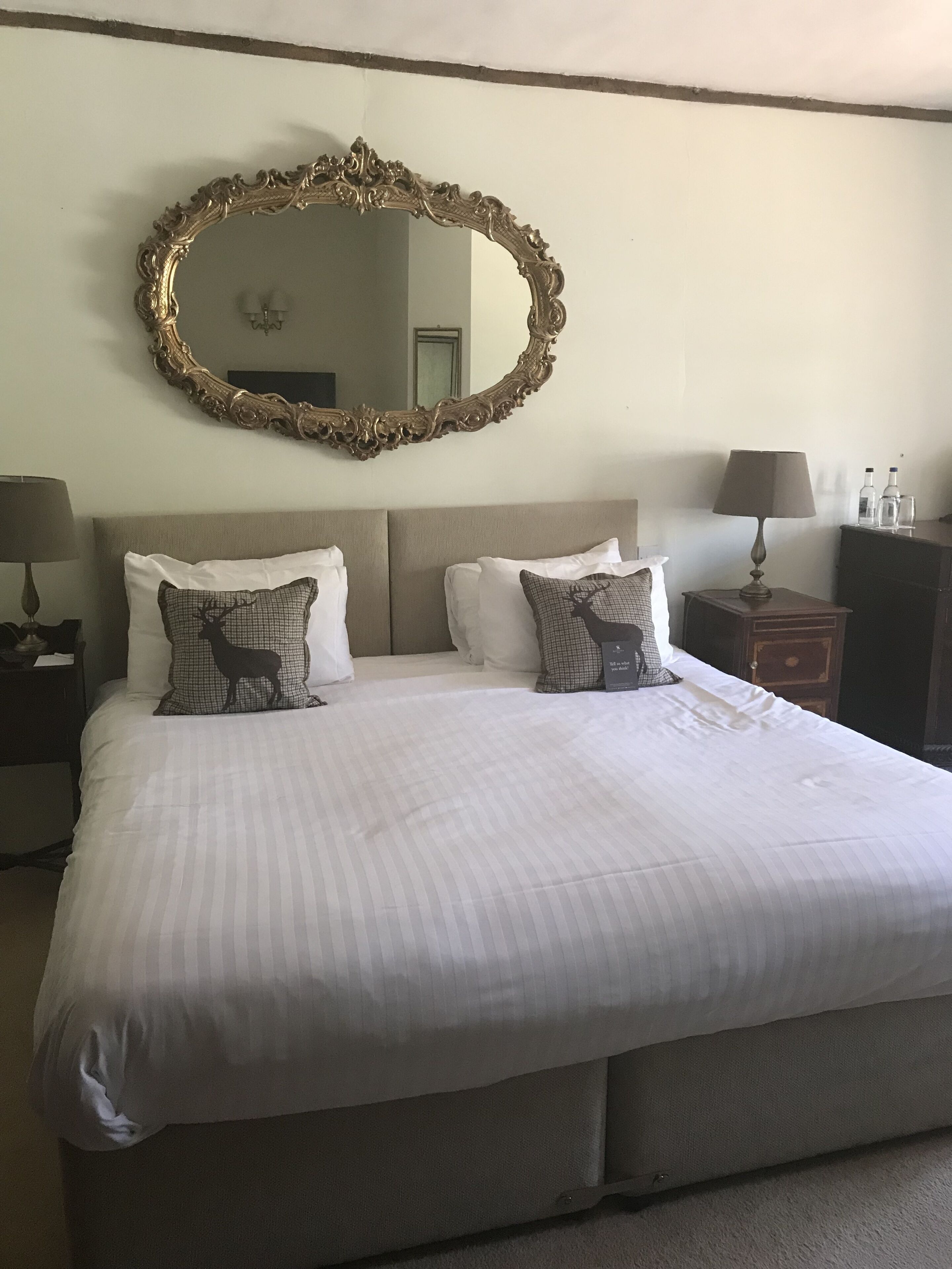 A neatly made bed with striped bedding, two bedside tables with lamps, a large ornate mirror, and a dresser with bottles and glasses.