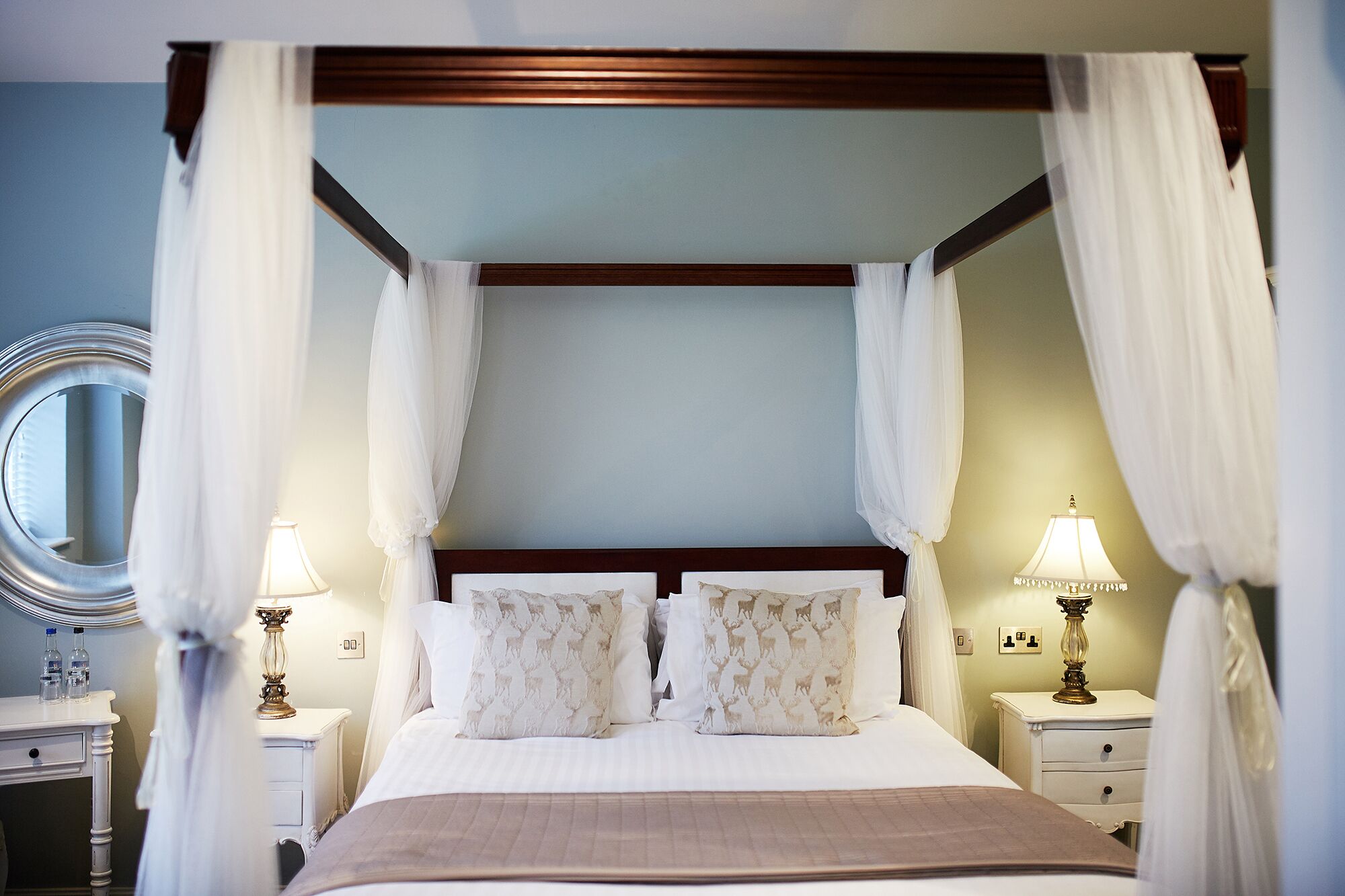 Un lit à baldaquin à quatre poteaux, agrémenté de draps blancs et d’oreillers à motifs, des tables de chevet avec des lampes et un miroir rond posé sur une commode.