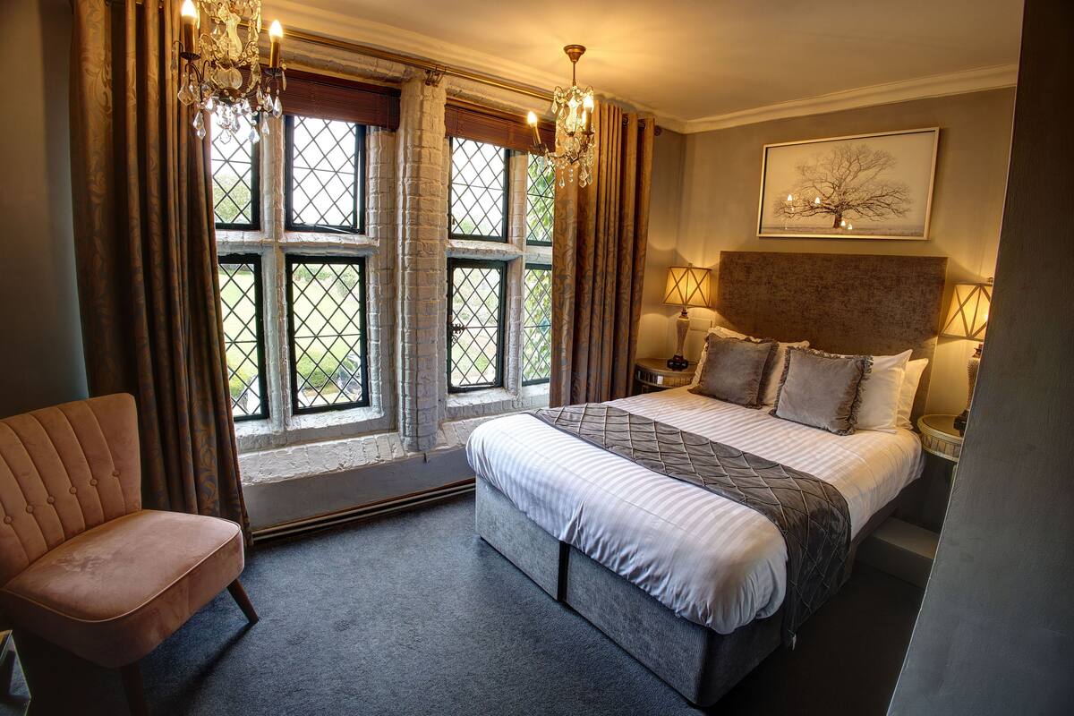 A bedroom with a large bed, a chair, a window with curtains, and a chandelier.
