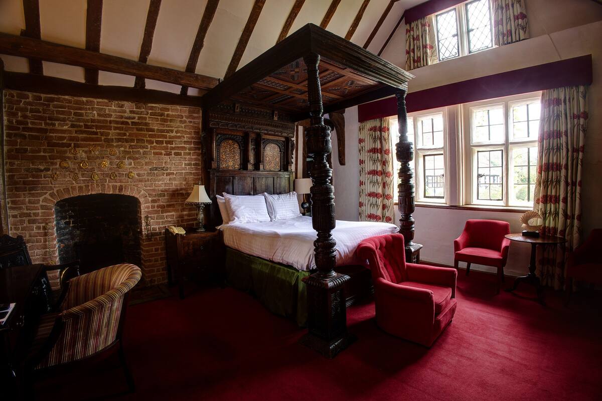 A traditional four-poster bed with a brick fireplace, red upholstered chairs, and a wooden table in a room with exposed wooden beams and large windows.