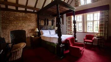 A traditional four-poster bed with a brick fireplace, red upholstered chairs, and a wooden table in a room with exposed wooden beams and large windows.