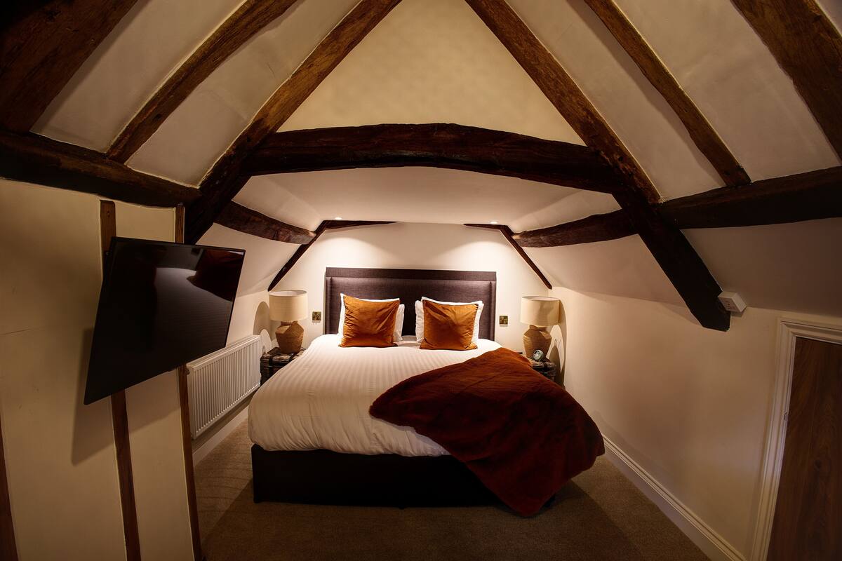 A bedroom with a sloped ceiling, a bed with white bedding and brown pillows, two bedside lamps, and a red throw blanket.