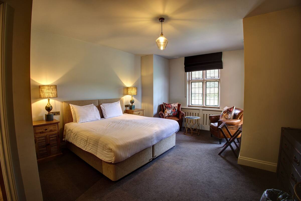 A bedroom with a large bed, two bedside tables with lamps, a chair, a small table, and a window with blinds.