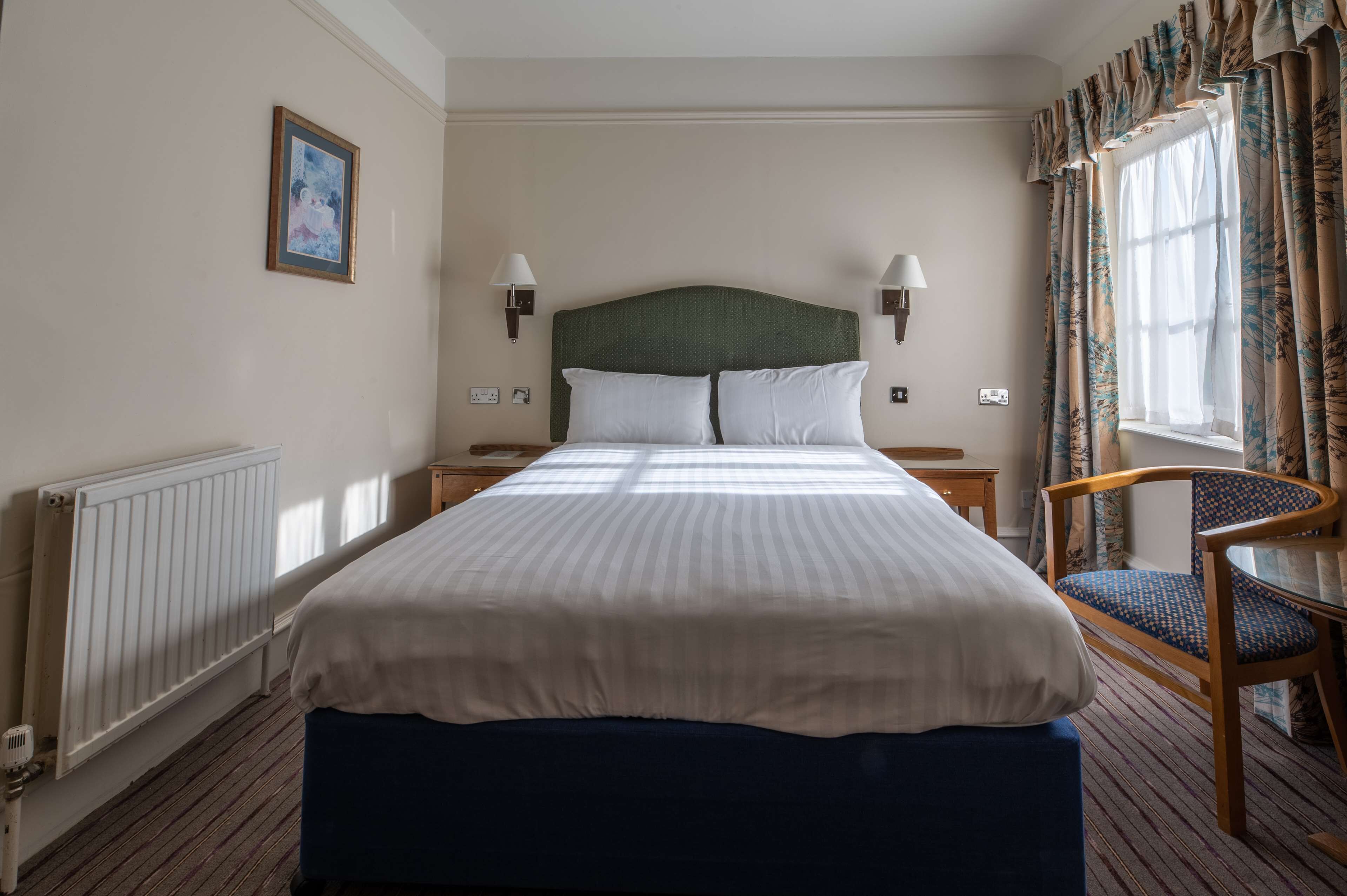Quarto de hotel com uma cama grande, duas mesas de cabeceira, uma cadeira, janela com cortinas e radiador.
