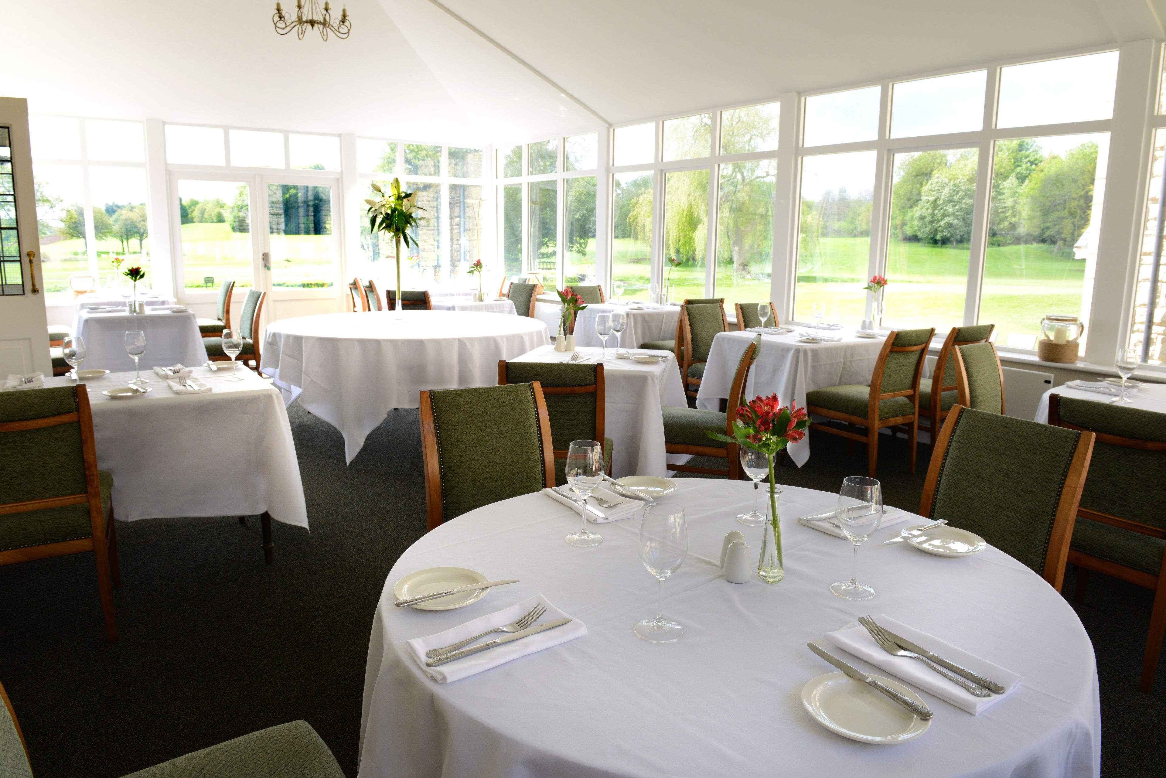A dining area with round tables set for four, each with white tablecloths, green upholstered chairs, and a vase with flowers.