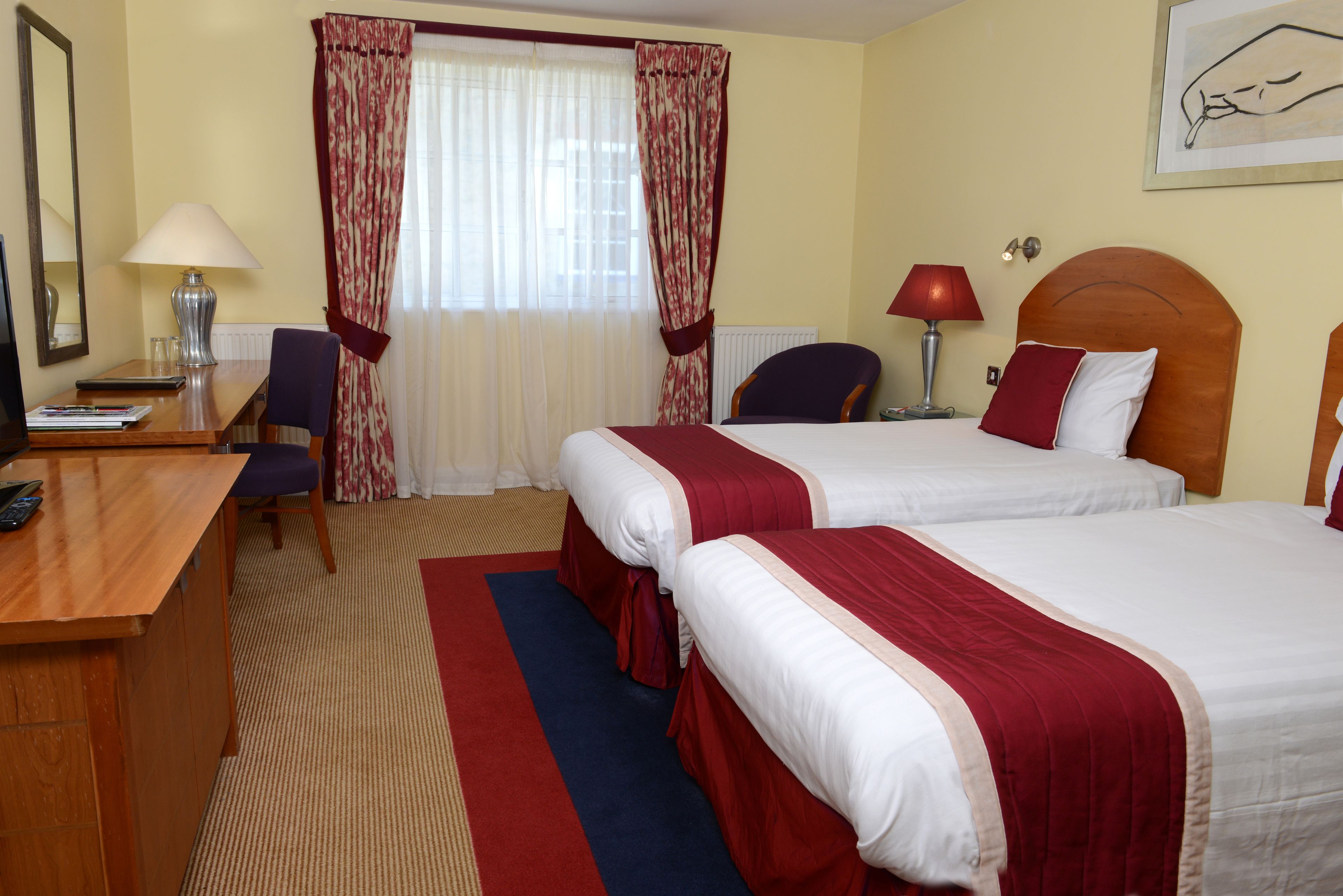 Une chambre d’hôtel avec deux lits, un bureau, une chaise, une lampe et une fenêtre avec des rideaux.