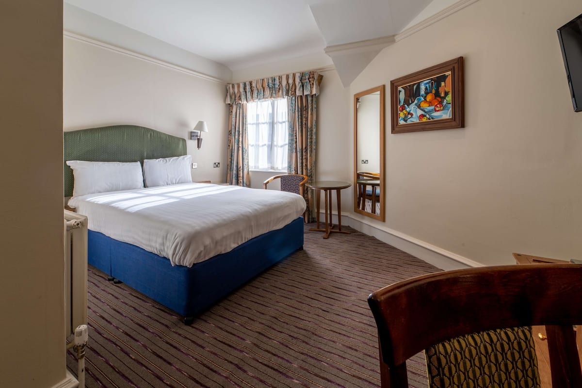 A hotel room with a large bed, a chair, a small table, and a painting on the wall.