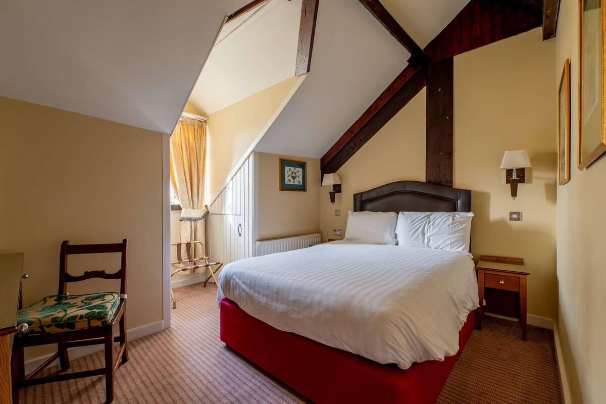 A bedroom with a large bed, a wooden chair, a nightstand, and a window with curtains.