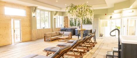 A spacious indoor area with wooden flooring, a sofa, lounge chairs, a hot tub, and a hanging plant.