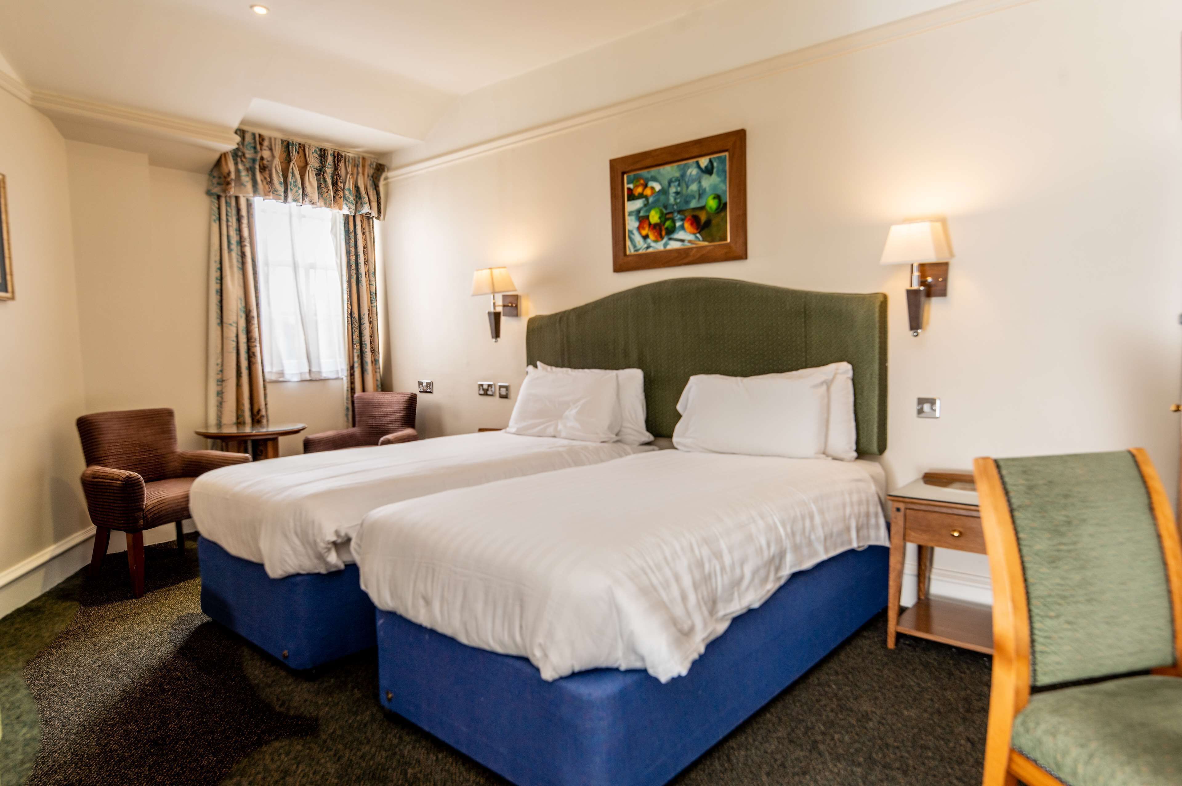 A hotel room with two beds, a small table, a chair, a lamp, and a painting on the wall.