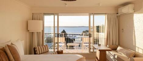 A hotel room with a large bed, a balcony with a view of the sea, a ceiling fan, and a sofa.