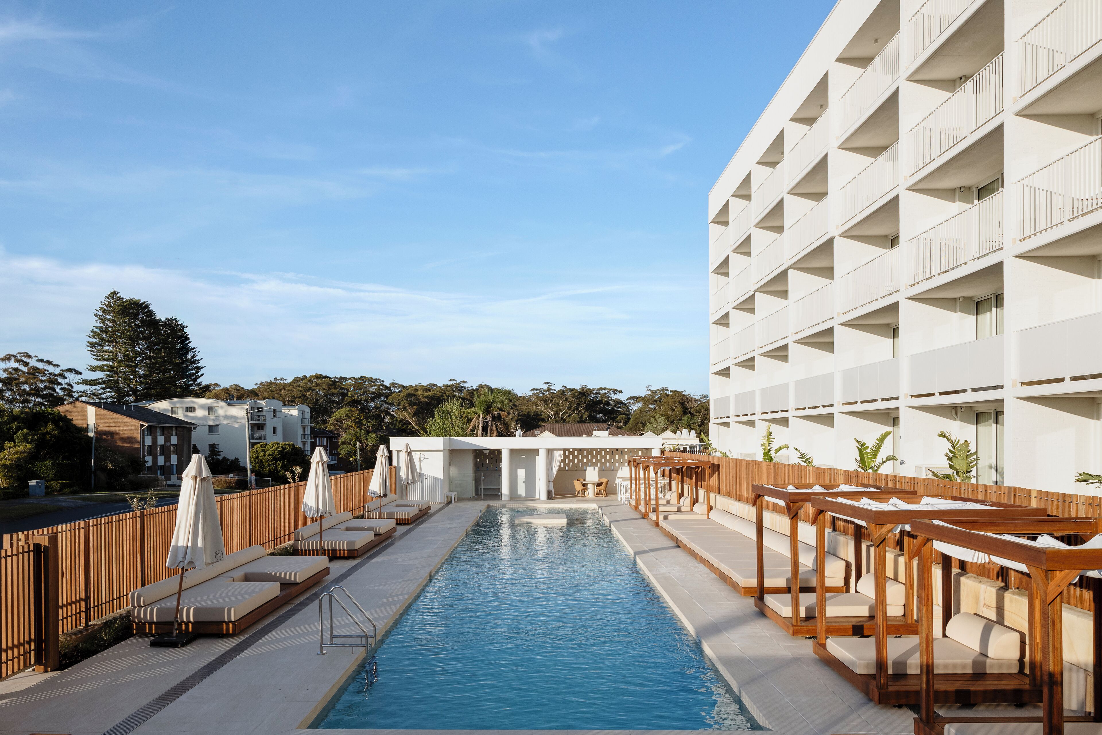 A pool area with lounge chairs and umbrellas, surrounded by a wooden fence, and adjacent to a white building with balconies.