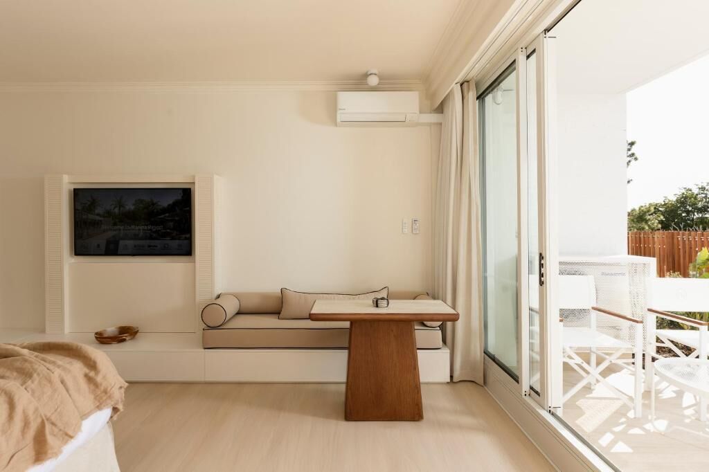 A modern living room with a sofa, a wooden coffee table, a flat-screen TV mounted on the wall, and a sliding glass door leading to an outdoor area with white furniture.