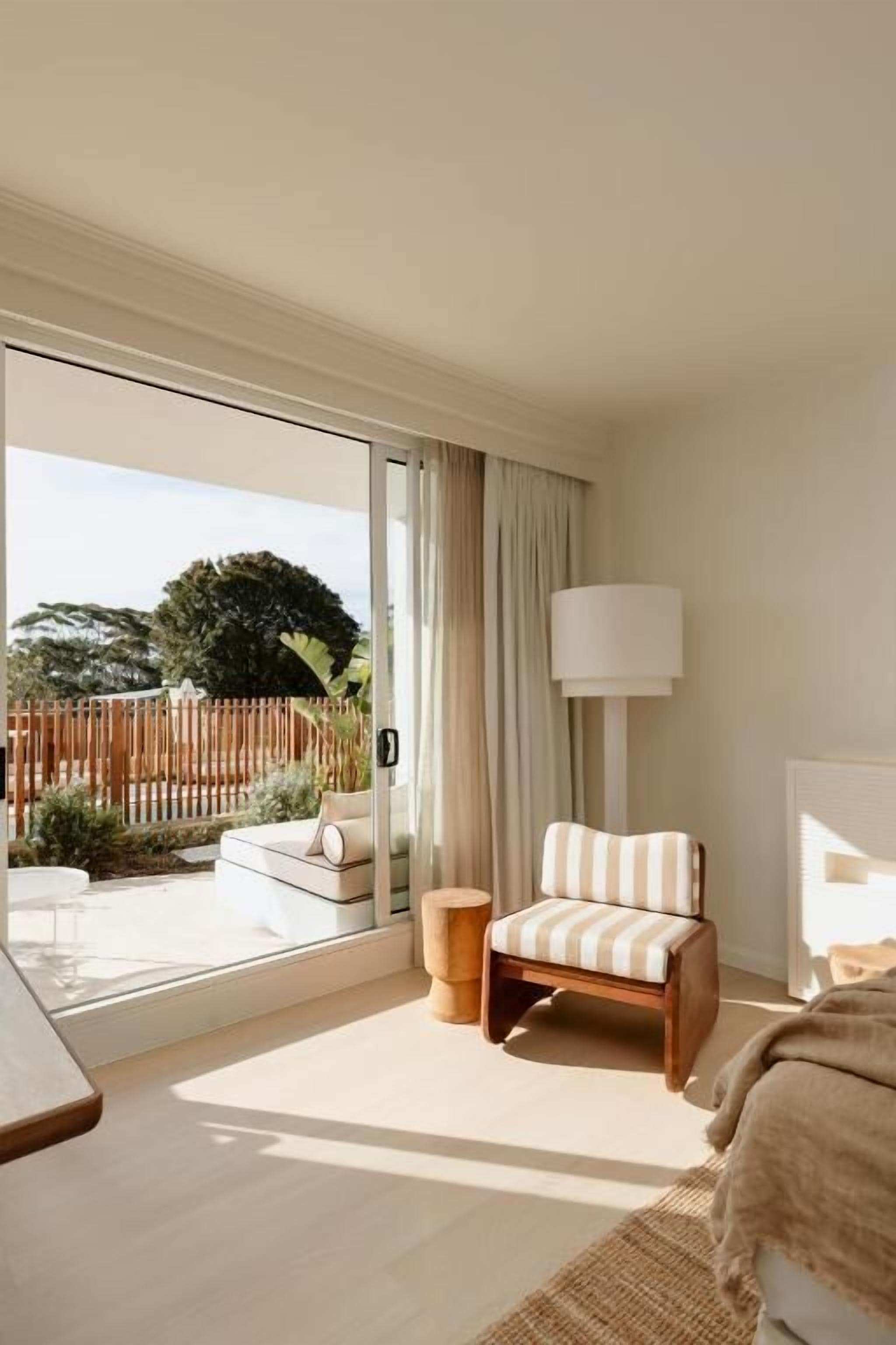 A hotel room with a large sliding glass door leading to a balcony, a striped armchair, a bedside table, and a lamp.