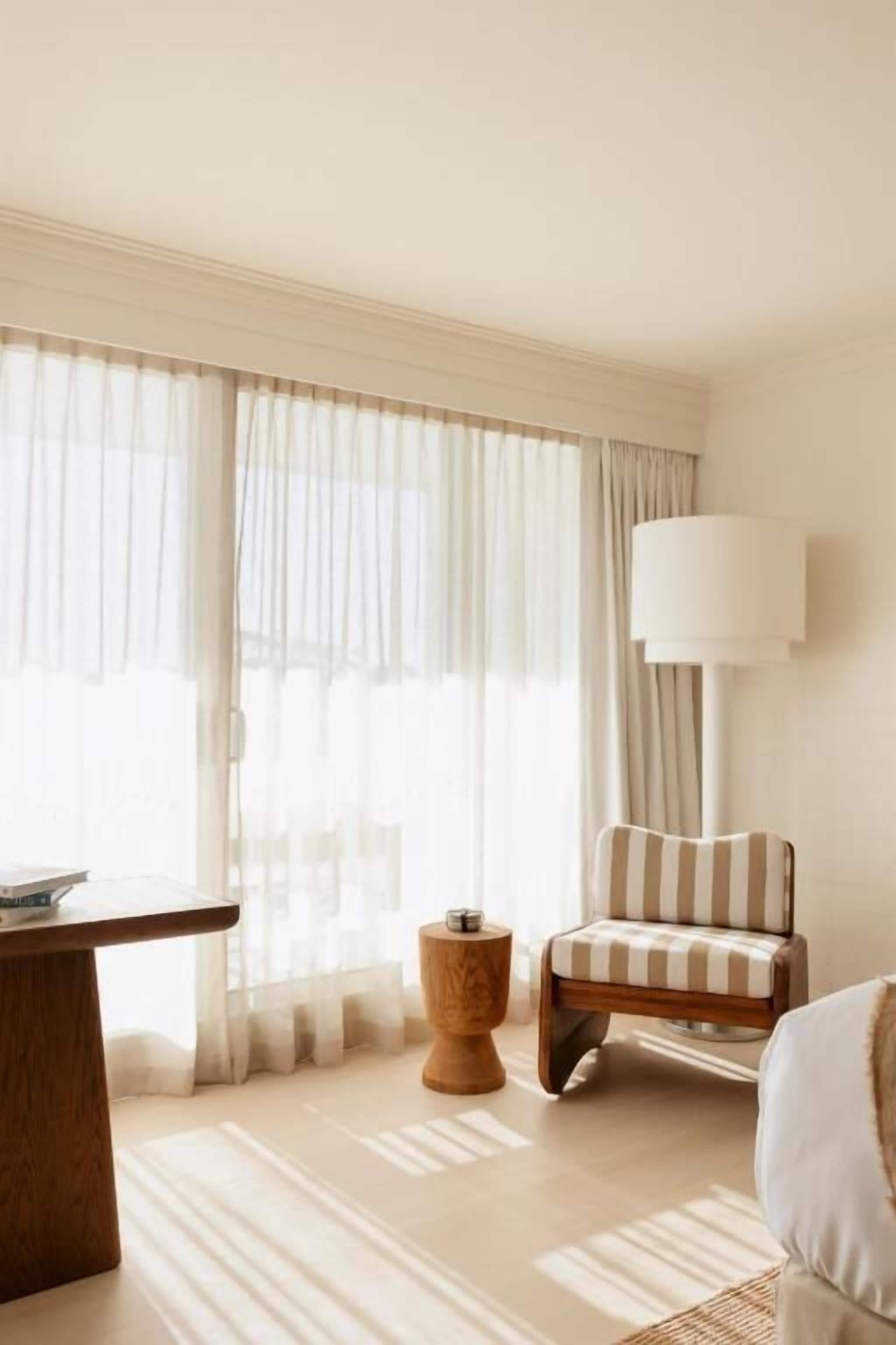 A hotel room with a large window, a wooden side table, a striped armchair, and a floor lamp.